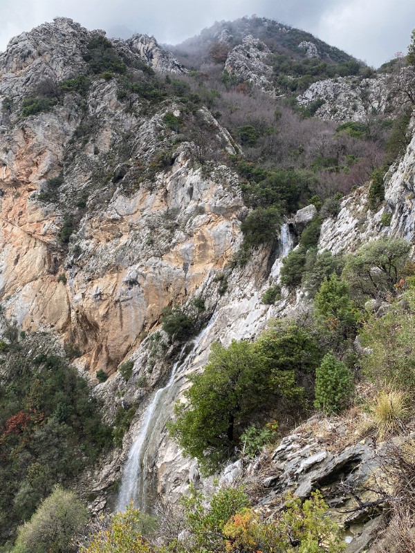 A photo of a tall, narrow waterfall cascading down a steep, rugged mountain face. The rocky cliffs are a mix of gray and light tan stone, with green shrubs, and small trees. The top of the mountain disappears into a misty, overcast gray sky.