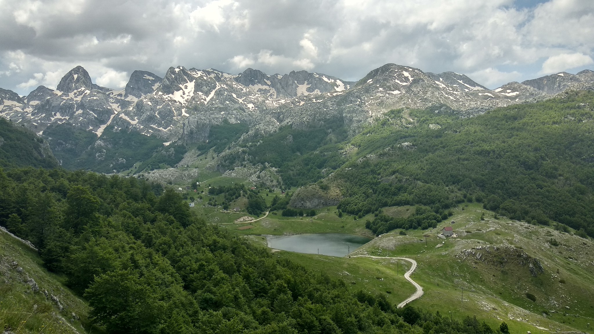 A wide panoramic photo of a mountain valley under a cloudy sky. In the background, a jagged, rocky mountain range stretches across the horizon, with patches of snow remaining on the peaks and slopes. Below the mountains, the landscape opens into a green valley filled with dense forests, grassy fields, a small round lake, and a winding dirt road leading through the valley floor.