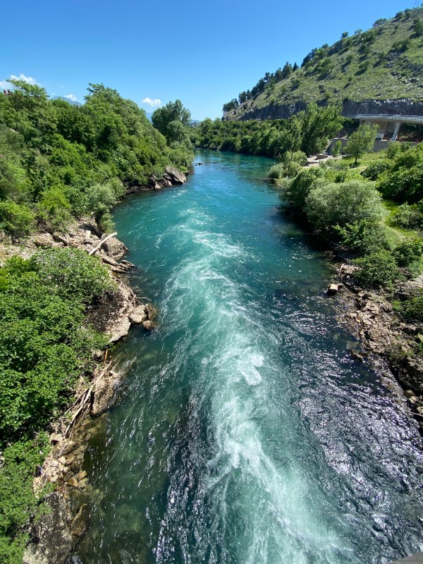 A photo of a rushing, turquoise-blue river cutting through a lush canyon. White rapids churn in the center of the current, flowing away from the foreground. The riverbanks are lined with dense green trees, rocky shores, and a steep hillside on the right, all under a clear, bright blue sky.