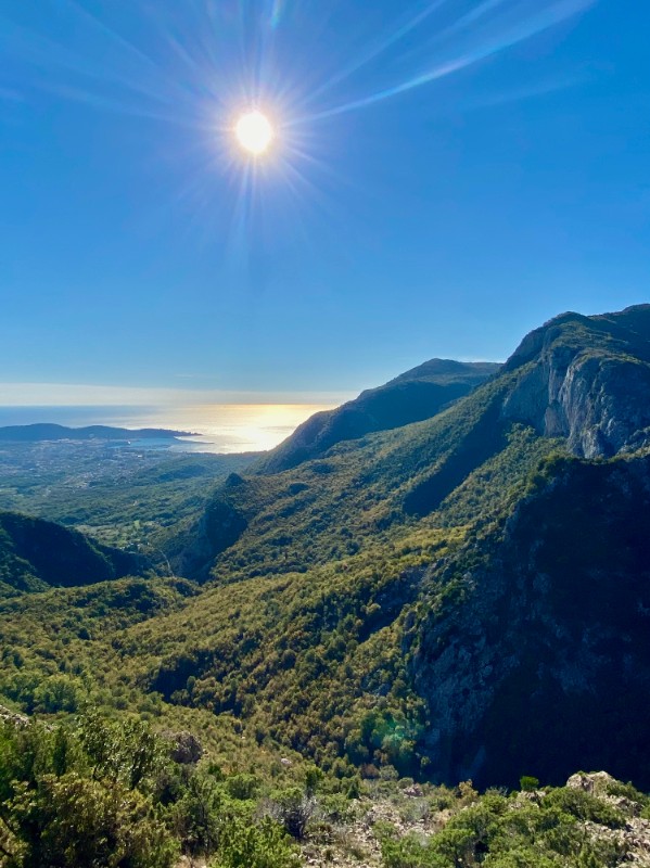 A bright, sunlit view from a high mountain looking down onto green forest and slopes. In the distance, the sun reflects brightly off the sea near a coastline and a small coastal town. The sun shines intensely in a clear blue sky, with a strong glare and long rays downward.