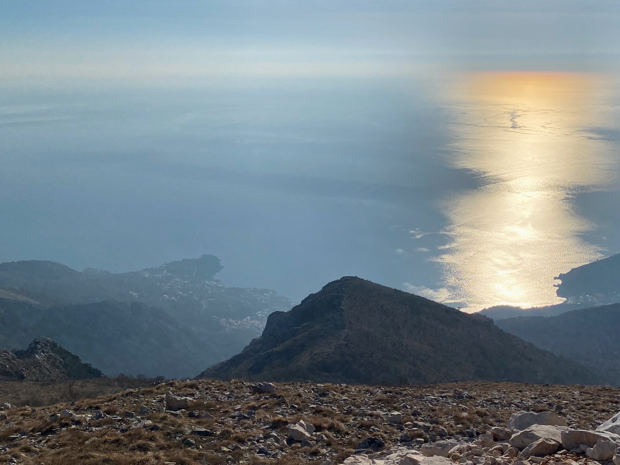 A high-angle view from a rocky mountain summit looking down at the sea. Bright golden sunlight reflects off the water on the right side of the frame. The ground is covered in rocks and dry grass. Below there&rsquo;s a mountain ridge that goes downward toward the coastline in the haze. A small coastal town is visible through the haze.