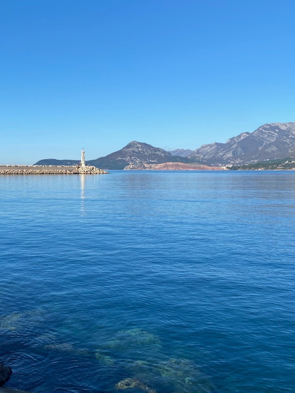 A small white lighthouse stands at the end of a stone breakwater. The sky is clear and blue. The sea is calm. There are mountains in the background.