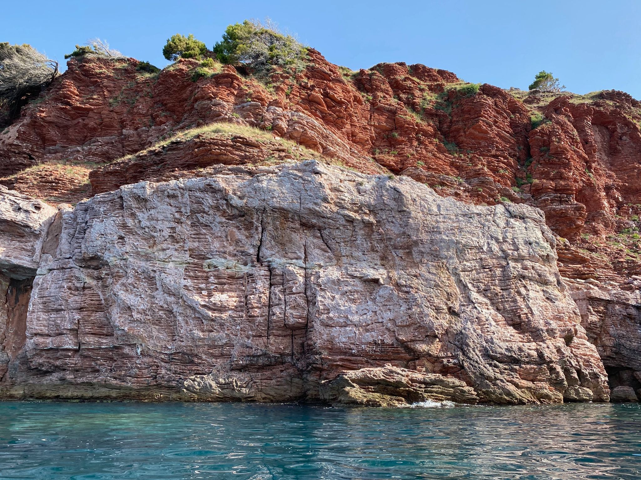 A photo of a coastal cliff rising directly from turquoise water under a clear blue sky. The rock formation is composed of geological layers: a large, light grey and pinkish limestone wall in the foreground, and a reddish-brown, layered sedimentary cliff directly behind it, with patches of green vegetation along the ridges.