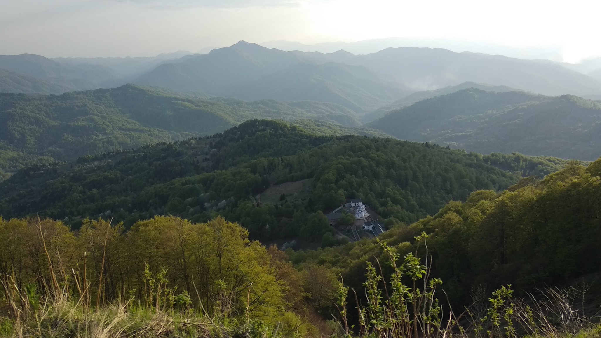High-angle view of mountain ranges stretching into the hazy background, with a small white monastery among dense trees on a hillside.