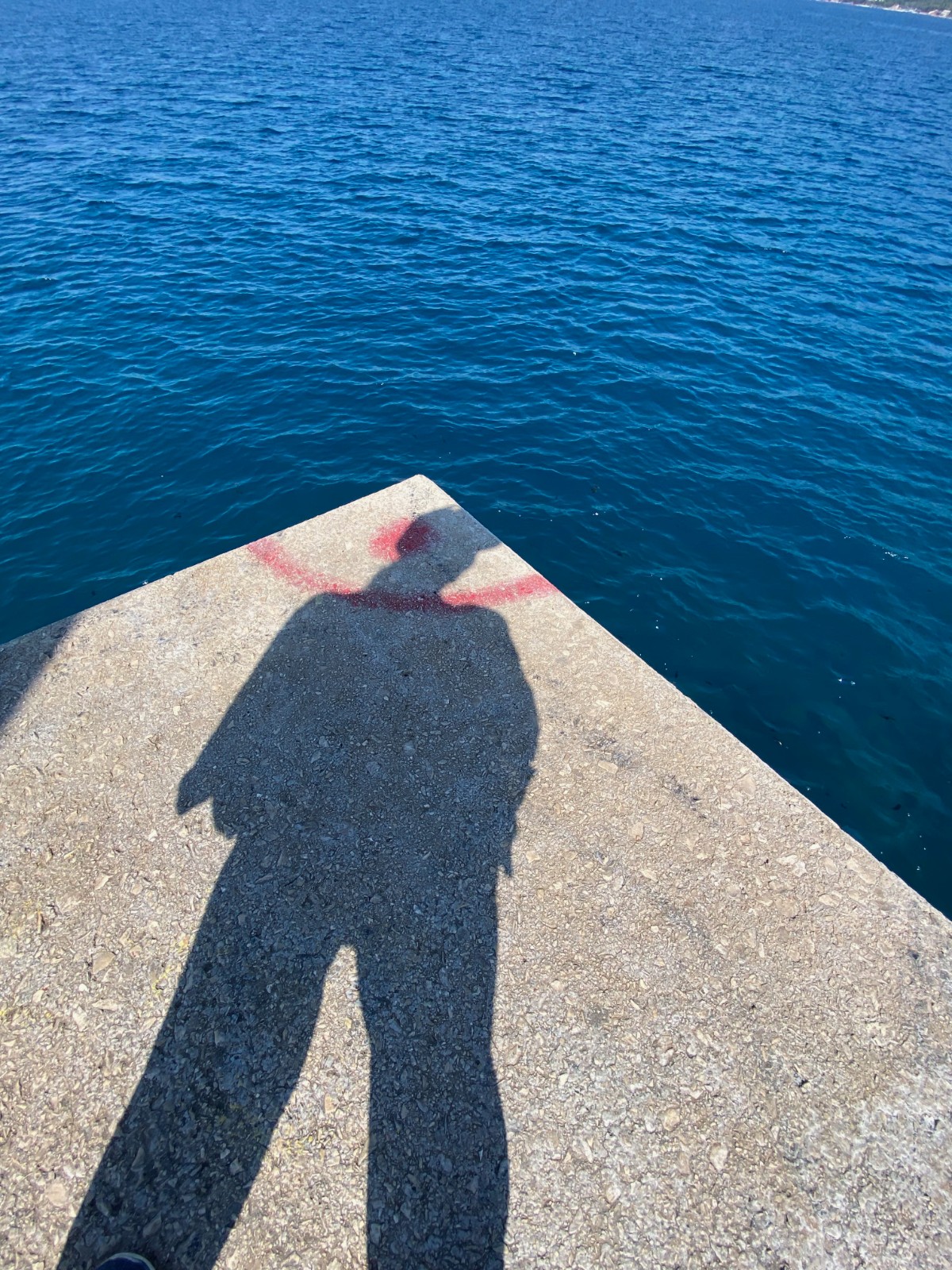A high-angle view of the shadow of a person on a concrete pier. The shadow is positioned over a red painted semi-circle, creating a halo effect. The edge of the pier forms a sharp corner, leading the eye to deep blue ocean water.