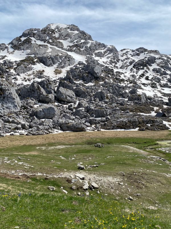 A photo showing a rugged limestone mountain partially covered in snow under a cloudy sky, with a grassy green valley containing a small stone picnic table in the foreground.