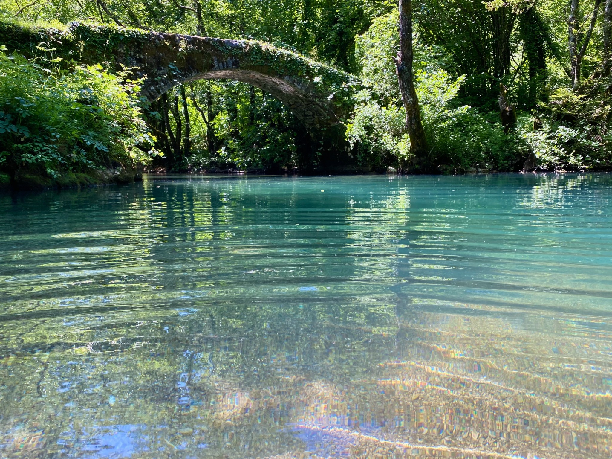 An old moss covered stone arch bridge over clear turquoise river in a lush green forest.